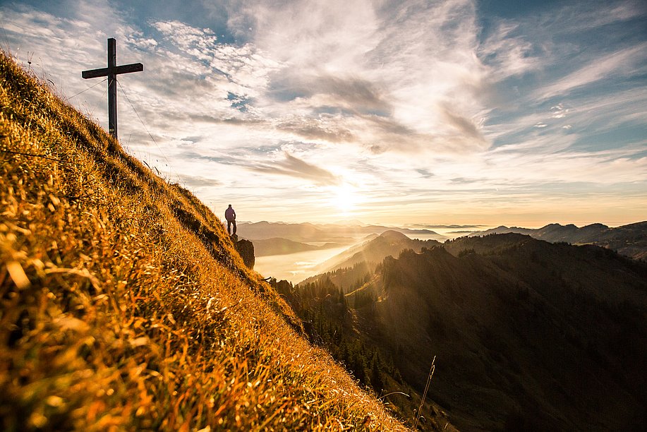 Gipfelkreuz des Siplinger Kopfs, daneben Mann, der in die Ferne über die Berge guckt