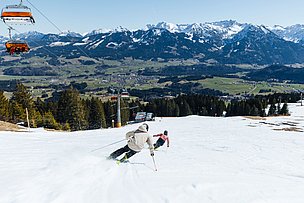 Zwei Skifahrer auf den Pisten der Hörnerbahn, es ist Frühjahr und das Tal ist schon grün, während die Gipfel noch weiß sind