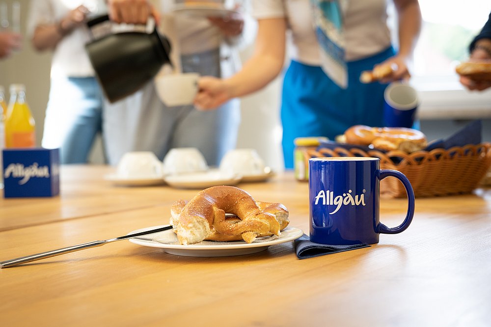 Allgäu-Tasse neben Teller mit Breze, im Hintergrund schenken Leute sich Kaffee ein