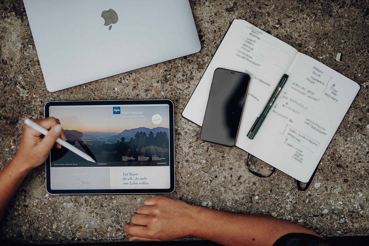 Ein Tablet, ein Notebook, ein Handy und ein Notizbuch auf einem Steintisch