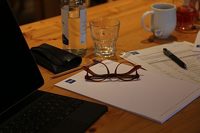 Laptop, Notizblock, Handy, Wasser, Kaffeetasse und Brille auf einem Holztisch