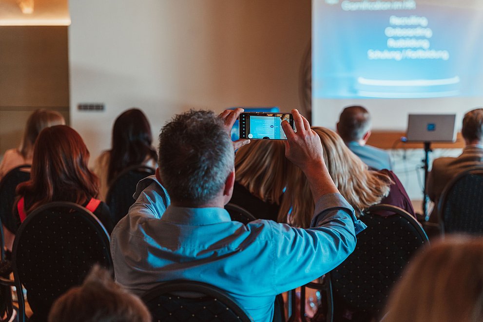 Publikum von hinten, Mann macht ein Handyfoto von einer Präsentation