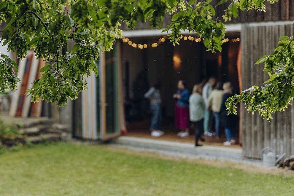 Im Vordergrund Äste mit Blättern, dahinter ein offenes Holzhaus mit Menschen, die sich unterhalten
