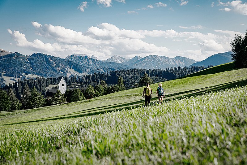 Zwei Wanderer auf einer Wiese, im Hintergrund ein Kirchturm vor Bergpanorama Zwei Wanderer auf einer Wiese, im Hintergrund ein Kirchturm vor Bergpanorama