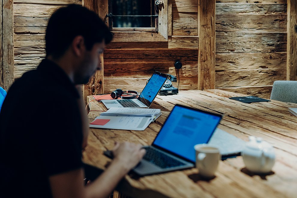 Mann an einem Laptop an einem Altholztisch, im Hintergrund ein weiterer Laptop mit Headset vor einer Altholzwand