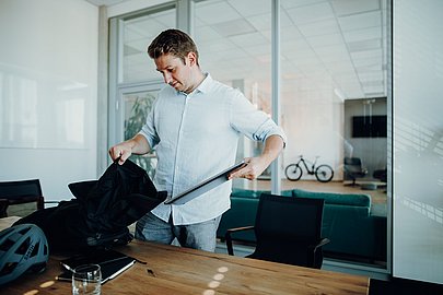 Mann in einem Büro packt seinen Laptopn in einen Rucksack, neben ihm ein Fahrradhelm auf dem Tisch