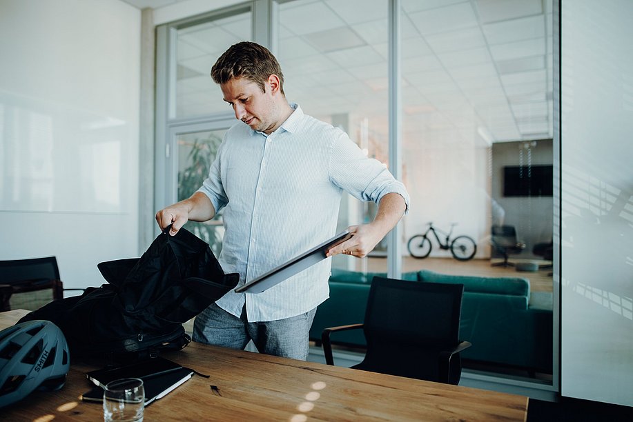 Mann in einem Büro packt seinen Laptopn in einen Rucksack, neben ihm ein Fahrradhelm auf dem Tisch